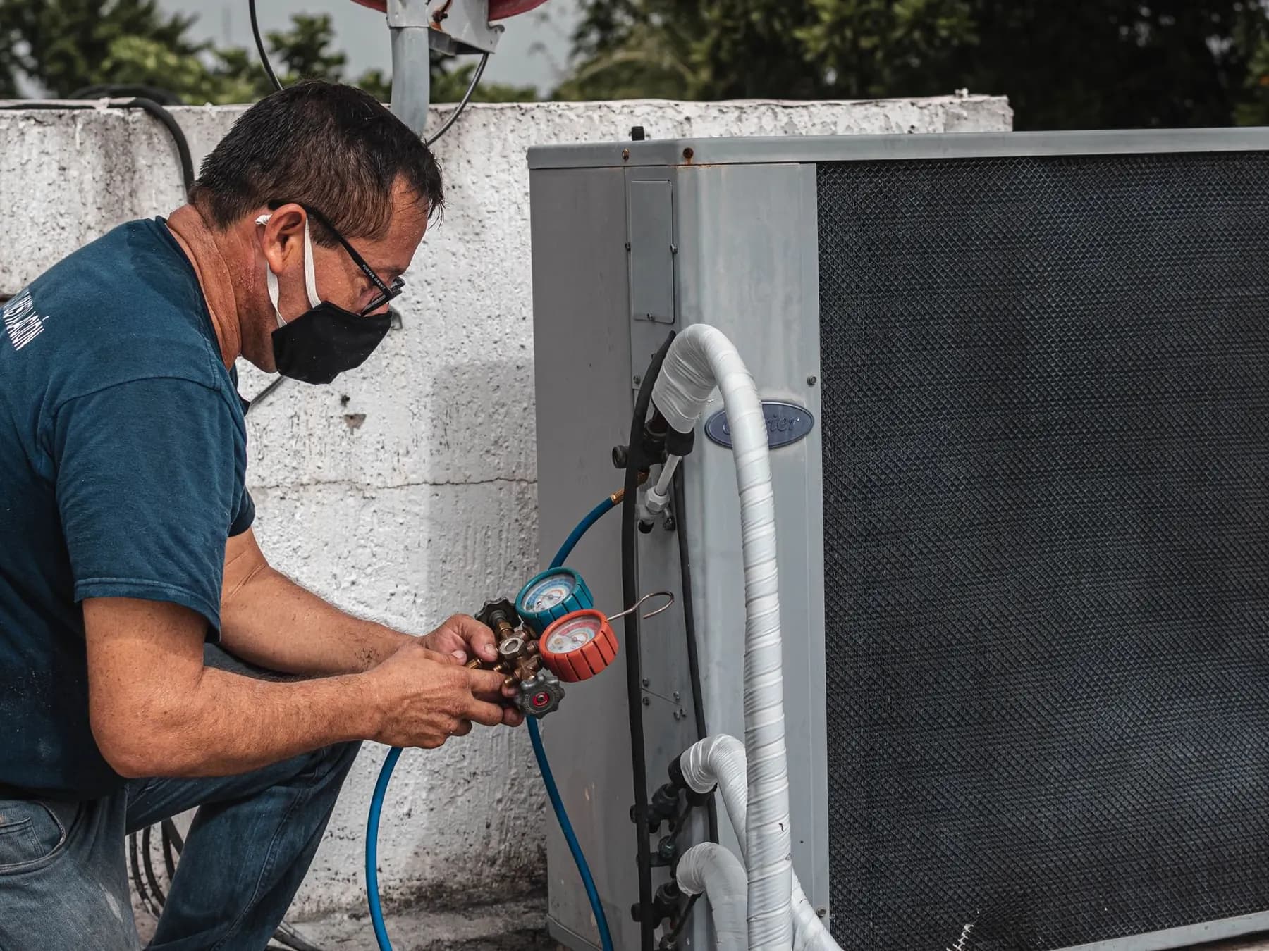 Technician crouched at an outdoor air conditioning condenser, holding a refrigerant manifold gauge set against the unit's service valves