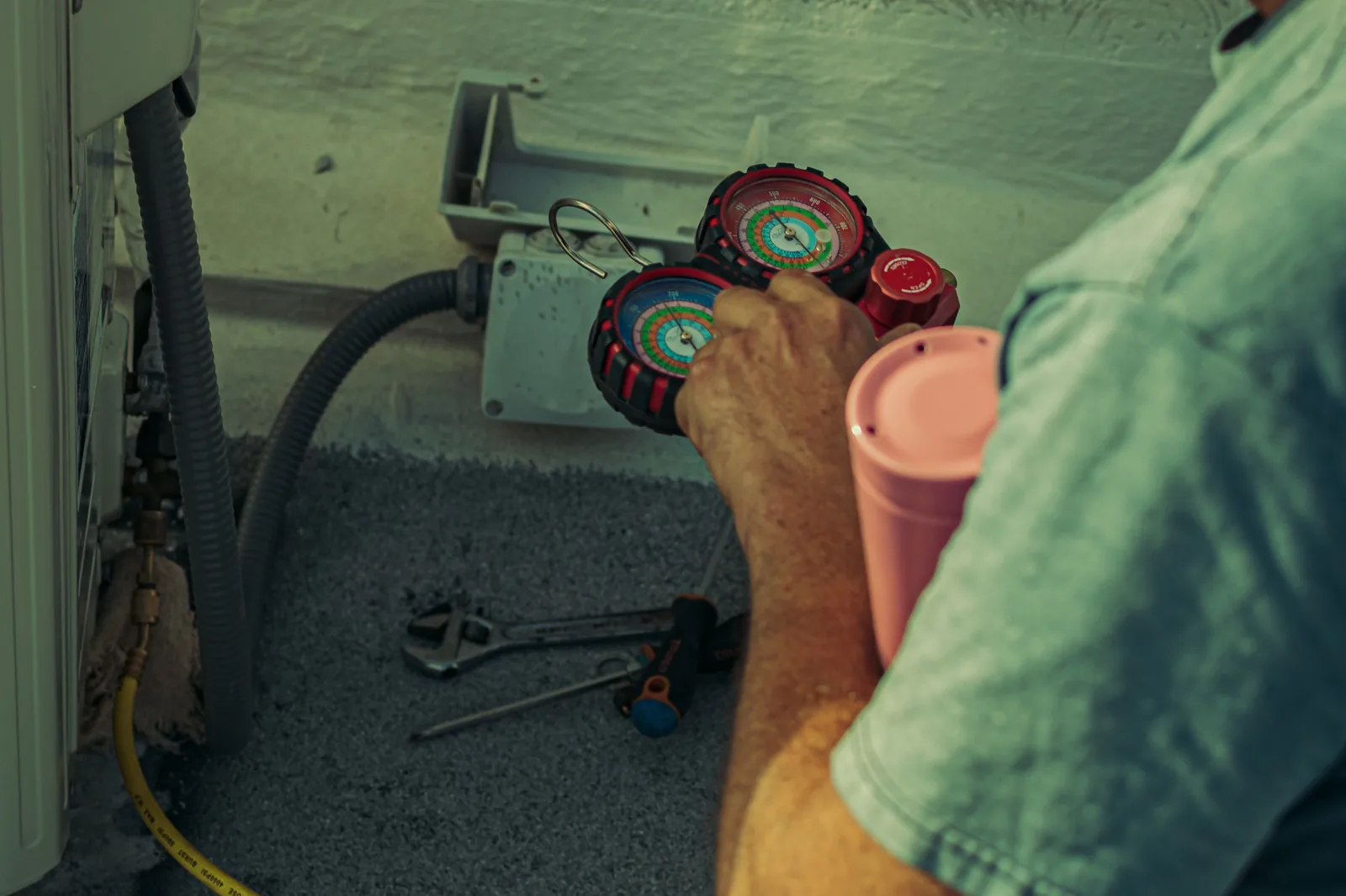 Refrigerant manifold gauges in a technician's hand at an outdoor air conditioning unit, with hand tools laid out alongside